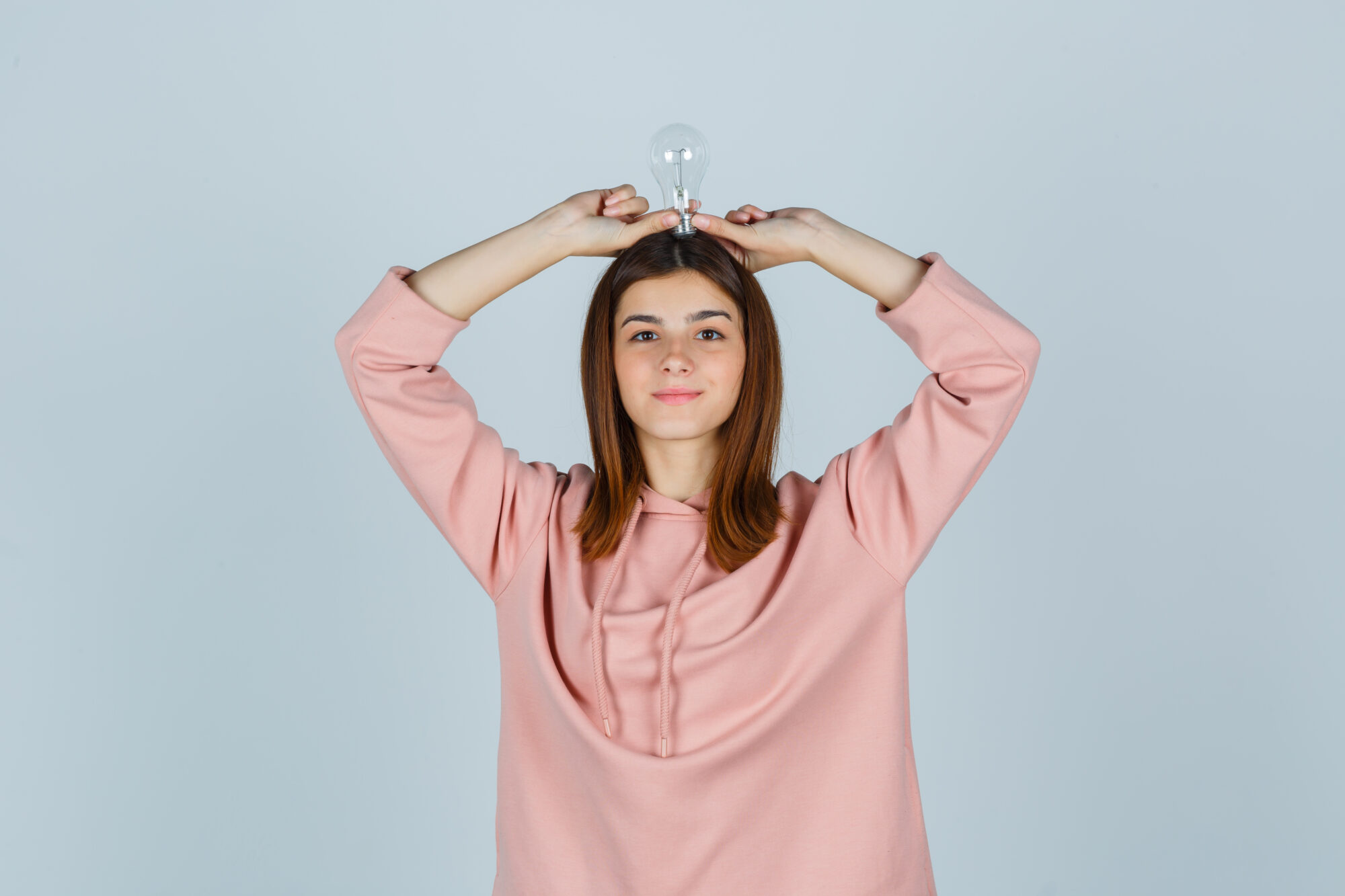 young lady holding light bulb over head in pink sweatshirt and looking pleased , front view.